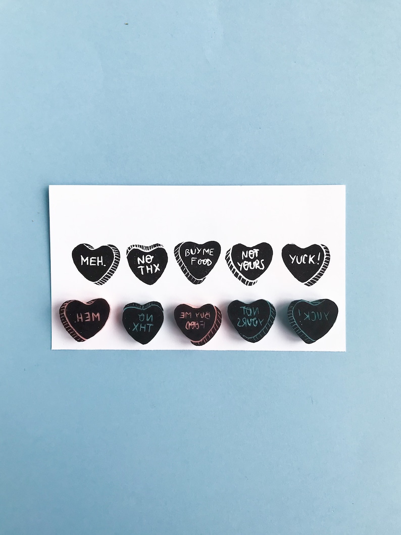 May include: A set of 10 black heart-shaped candy conversation hearts with various messages written in white, pink, and blue ink. The messages include "MEH", "NO THX", "BUY ME FOOD", "NOT YOURS", "YUCK!" and their reversed versions.