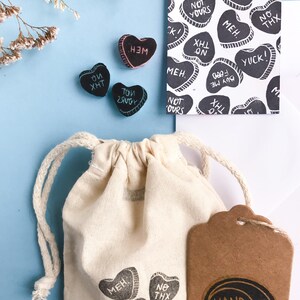 May include: A set of small, heart-shaped rubber stamps with various phrases, including "Not Yours", "Meh", "No Thx", and "Buy Me Food". The stamps are packaged in a small white drawstring bag with a brown paper tag that says "Hand Made".