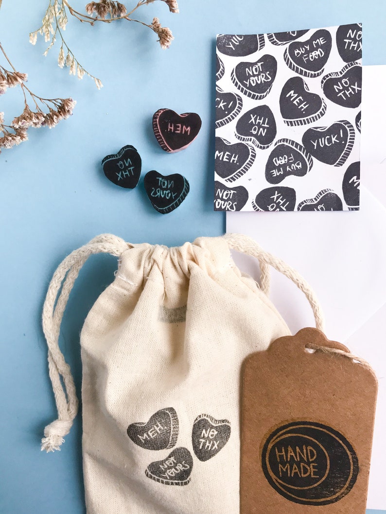 May include: A set of small, heart-shaped rubber stamps with various phrases, including "Not Yours", "Meh", "No Thx", and "Buy Me Food". The stamps are packaged in a small white drawstring bag with a brown paper tag that says "Hand Made".