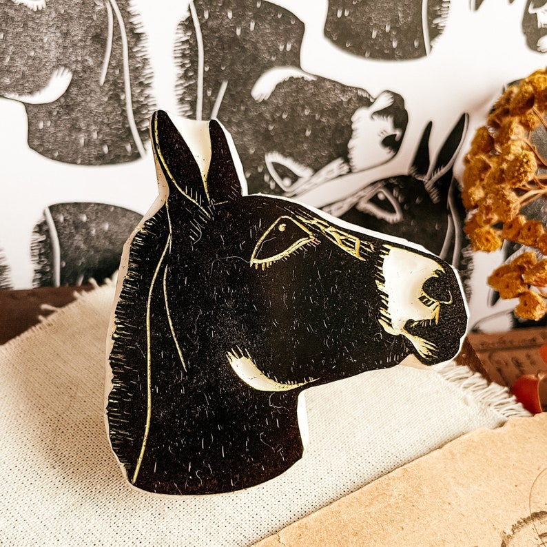 May include: A black and white donkey head art piece. The donkey's head is carved with a detailed design, featuring a white muzzle and ear tips. The background includes a print of donkeys and dried flowers, creating a rustic aesthetic.
