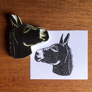 May include: A black and white donkey head linocut print and the linoleum block used to create it. The donkey is facing left, with detailed features. The linoleum block is black with yellow highlights. The print is on white paper.