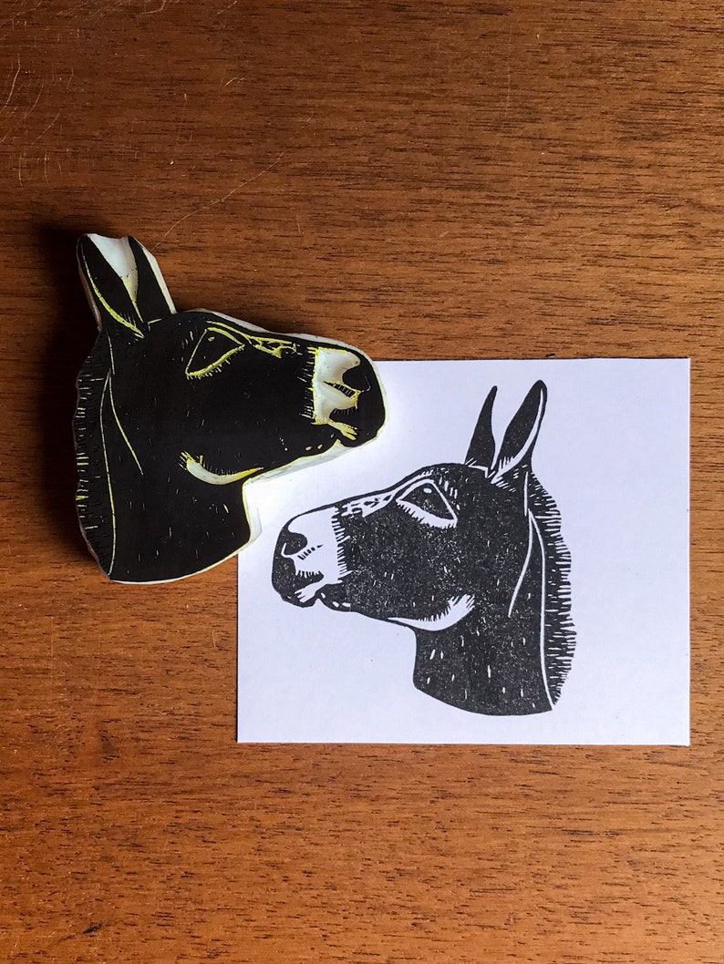 May include: A black and white donkey head linocut print and the linoleum block used to create it. The donkey is facing left, with detailed features. The linoleum block is black with yellow highlights. The print is on white paper.