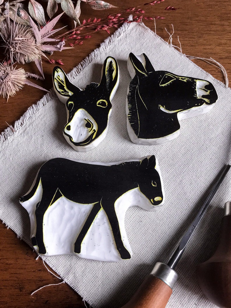 May include: Three black and white donkey linocut prints. One shows a full donkey, and two show the donkey's head. The prints are on a white surface with yellow accents. Carving tools are in the background.