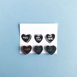 May include: Six black heart-shaped candies with white text. The text on the candies reads "MEH.", "NO THX", "NOT YOURS", "НЕМ", "ОЙ ХНТ", and "ТОИ ЗЯУОУ".
