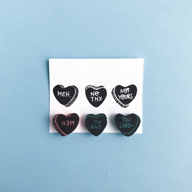 May include: Six black heart-shaped candies with white text. The text on the candies reads "MEH.", "NO THX", "NOT YOURS", "НЕМ", "ОЙ ХНТ", and "ТОИ ЗЯУОУ".