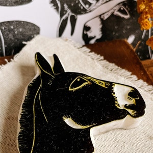 May include: A black and white donkey head sculpture with gold accents. The donkey head is carved from a light-colored material and sits on a beige cloth. In the background, there is a black and white donkey print.