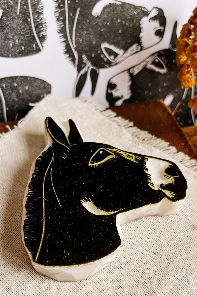 May include: A black and white donkey head sculpture with gold accents. The donkey head is carved from a light-colored material and sits on a beige cloth. In the background, there is a black and white donkey print.