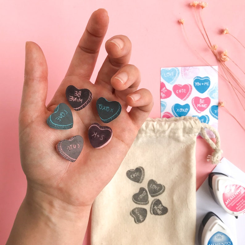 May include: A hand holding six small wooden heart-shaped stamps with different messages, including "Love", "Be Mine", "Cute", "XOXO", and "You + Me". The stamps are on a pink background with a white bag and a sheet of stickers with similar messages.