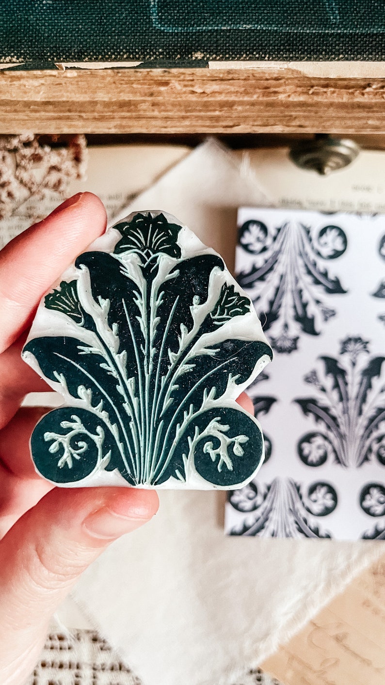May include: A green and white rubber stamp with a floral design. The stamp is shaped like a fan and has a detailed design of leaves and flowers.