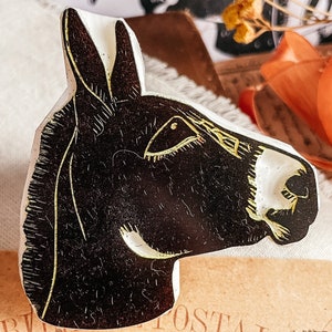 May include: A black and white donkey head-shaped decorative object with gold accents. The donkey's head is in profile, with detailed linework. The object is likely a stamp or a decorative element, possibly for crafts or art projects.