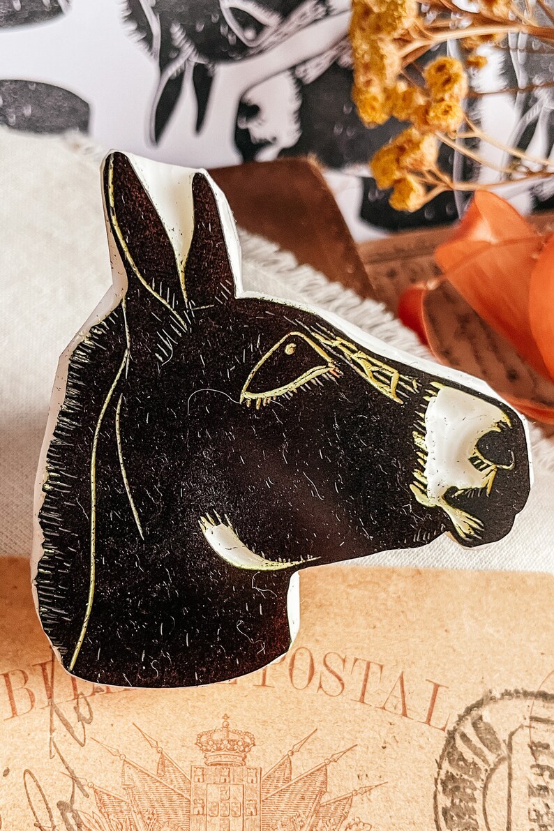 May include: A black and white donkey head-shaped decorative object with gold accents. The donkey's head is in profile, with detailed linework. The object is likely a stamp or a decorative element, possibly for crafts or art projects.