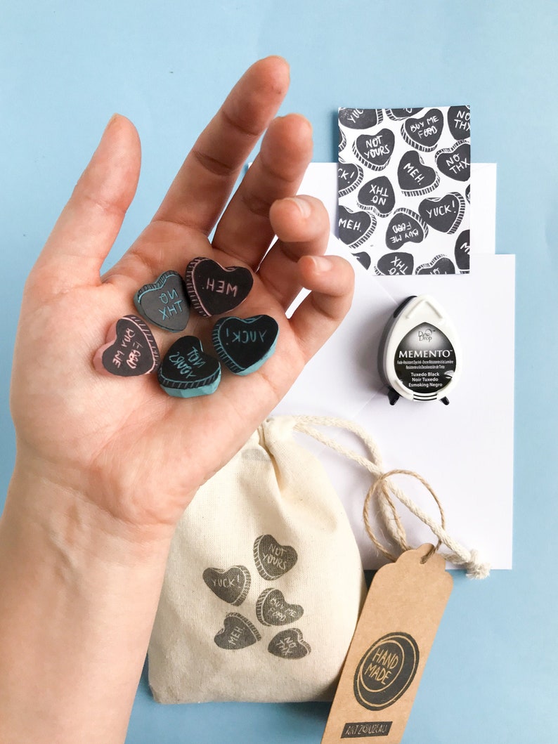 May include: A set of six heart-shaped rubber stamps with various phrases, including "YOU XHT", "MEH", "I  JUY", "BUY ME FORD", and "YUCK!" The stamps are in a hand, and there is a small bag with the stamps printed on it. The bag has a brown tag that says "HAND MADE" in a circle.