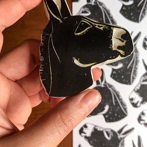 May include: A black and white donkey head stamp held by a hand, with a matching print in the background. The stamp has a carved design with yellow accents, showcasing the donkey's features. The print shows multiple donkey heads.