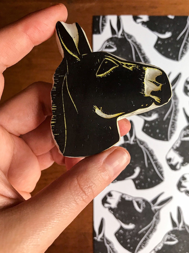 May include: A black and white donkey head stamp held by a hand, with a matching print in the background. The stamp has a carved design with yellow accents, showcasing the donkey's features. The print shows multiple donkey heads.