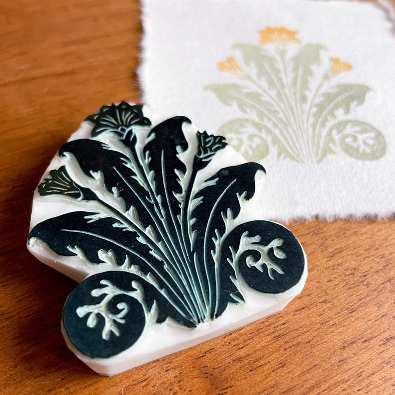 May include: A white rubber stamp with a black and green design of a stylized floral motif. The stamp is on a wooden surface.