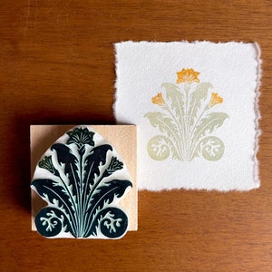 May include: A green and white rubber stamp with a floral design. The stamp is mounted on a wooden block. The design features a stylized flower with leaves and a central bud.