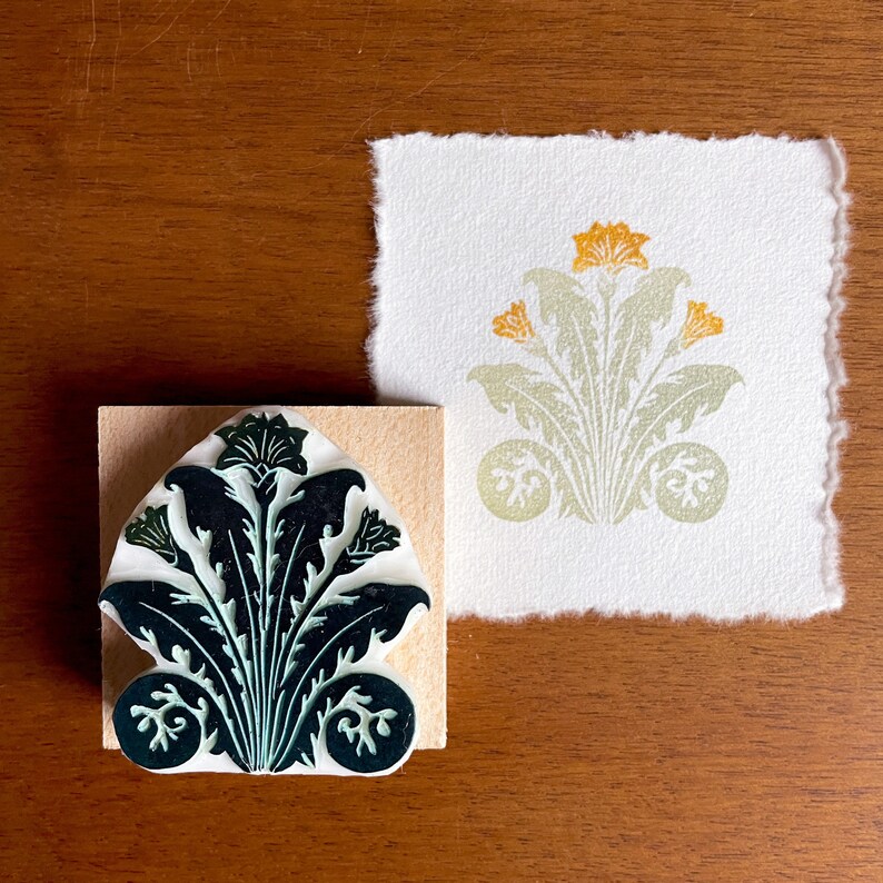 May include: A green and white rubber stamp with a floral design. The stamp is mounted on a wooden block. The design features a stylized flower with leaves and a central bud.