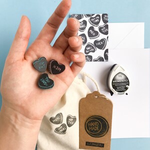 May include: A set of four black heart-shaped stamps with different phrases on them, including "Meh", "Not Yours", "To You", and "No Thx". The stamps are arranged on a hand, with a white bag and a brown tag that says "Hand Made" in the background.