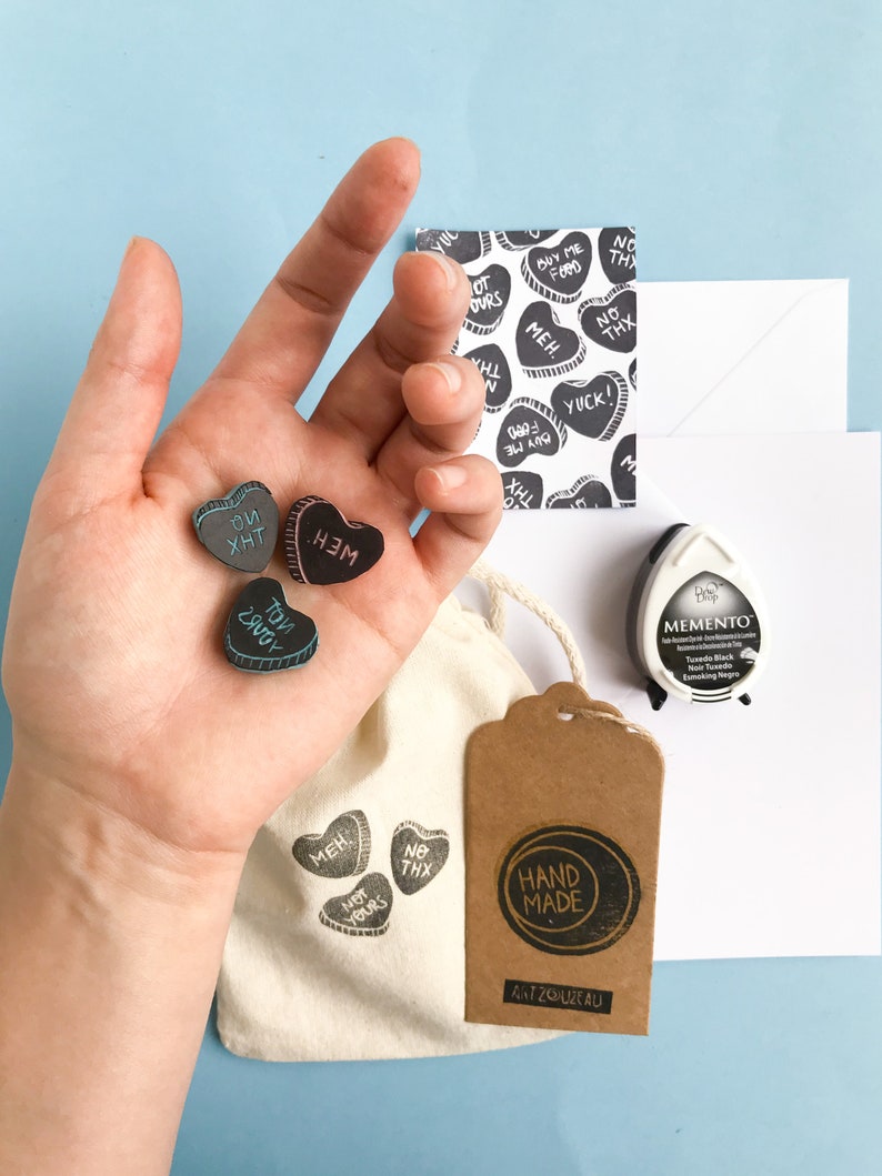 May include: A set of four black heart-shaped stamps with different phrases on them, including "Meh", "Not Yours", "To You", and "No Thx". The stamps are arranged on a hand, with a white bag and a brown tag that says "Hand Made" in the background.