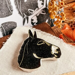 May include: A black and white donkey head-shaped ceramic art piece. The donkey's head is facing right, with a white muzzle and yellow accents. Two black and white donkey head prints are in the background, along with dried flowers and fabric.