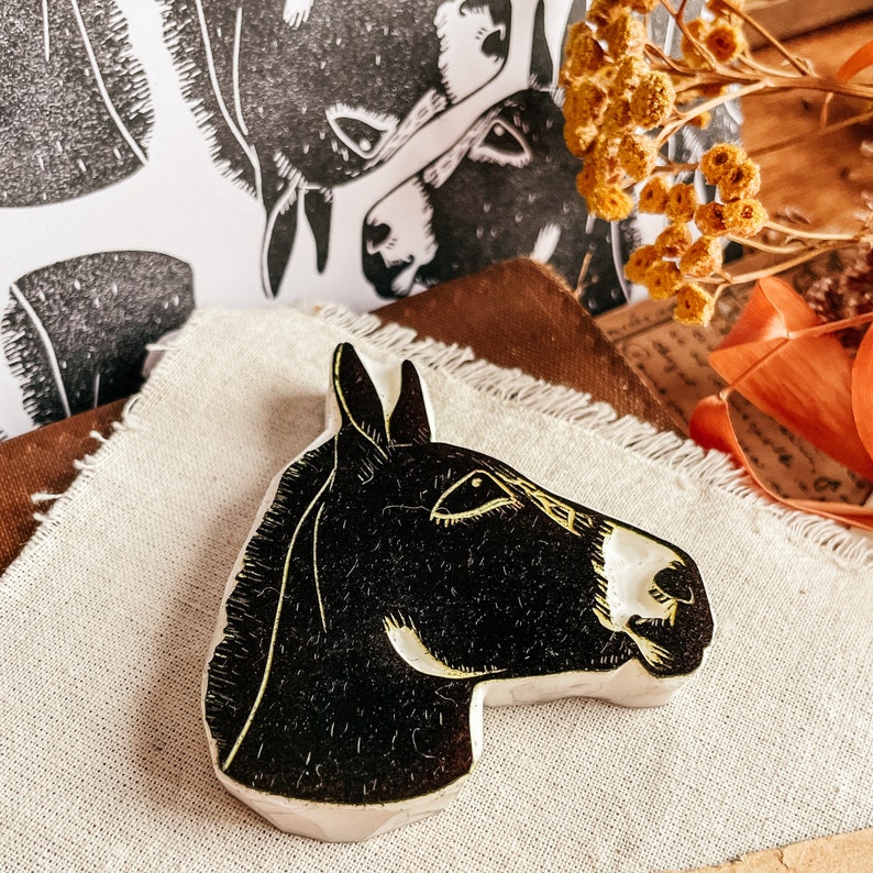 May include: A black and white donkey head-shaped ceramic art piece. The donkey's head is facing right, with a white muzzle and yellow accents. Two black and white donkey head prints are in the background, along with dried flowers and fabric.