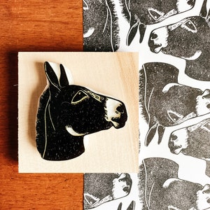 May include: A black and white donkey head woodcut print and a wooden block with a donkey head carved into it. The donkey head is in profile, with a white muzzle and ear. The print has a repeating pattern of donkey heads.