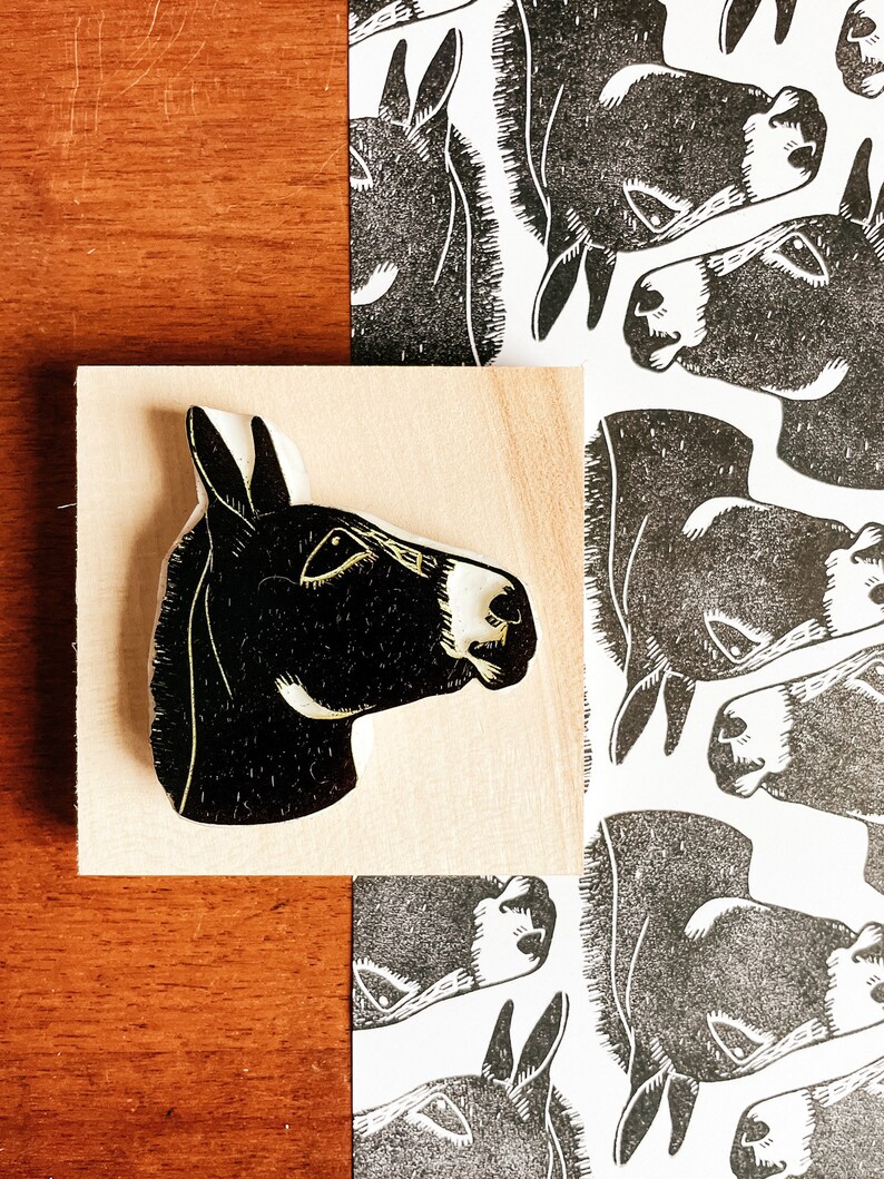 May include: A black and white donkey head woodcut print and a wooden block with a donkey head carved into it. The donkey head is in profile, with a white muzzle and ear. The print has a repeating pattern of donkey heads.