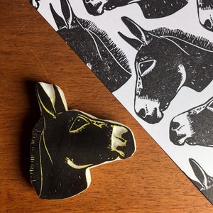 May include: A black and white donkey head print and a donkey head stamp. The stamp is black with yellow accents. The print features multiple donkey head illustrations on a white background. The items are on a wooden surface.