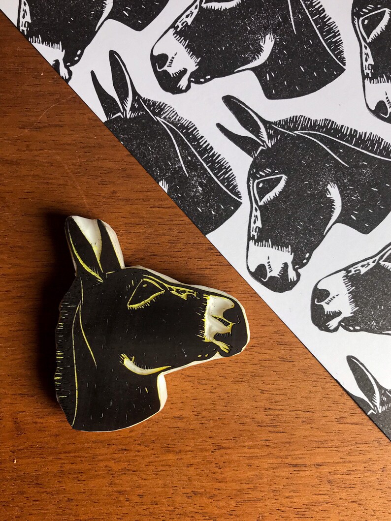 May include: A black and white donkey head print and a donkey head stamp. The stamp is black with yellow accents. The print features multiple donkey head illustrations on a white background. The items are on a wooden surface.