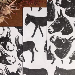 May include: Black and white donkey print on white paper. The image features multiple prints of donkeys in various poses, including full body and headshots. The prints are arranged on a wooden surface with dried plants in the background.