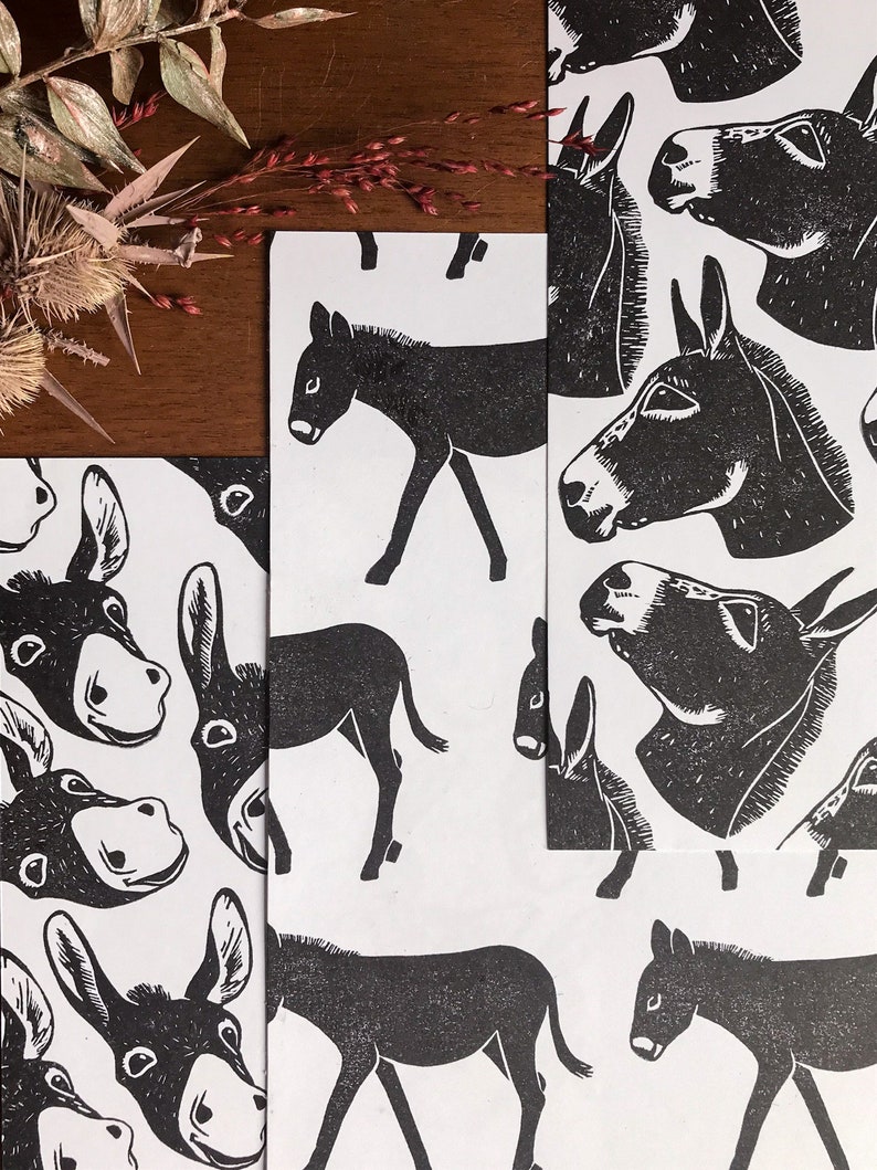 May include: Black and white donkey print on white paper. The image features multiple prints of donkeys in various poses, including full body and headshots. The prints are arranged on a wooden surface with dried plants in the background.