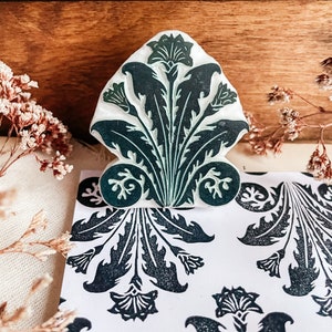 May include: A green and white rubber stamp with a floral design. The stamp is shaped like a leaf and has a detailed pattern of leaves and flowers. The stamp is sitting on a white surface with a black and white floral pattern.