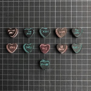 May include: A collection of 12 heart-shaped candy conversation hearts with various messages written on them, including "YOU + ME", "LOVE", "BE MINE", "XOXO", "YUCK", "THX", "CUTE", "TOU + ME", "YOU'RE", and "TOU + ME". The hearts are arranged in three rows of four on a black grid background.