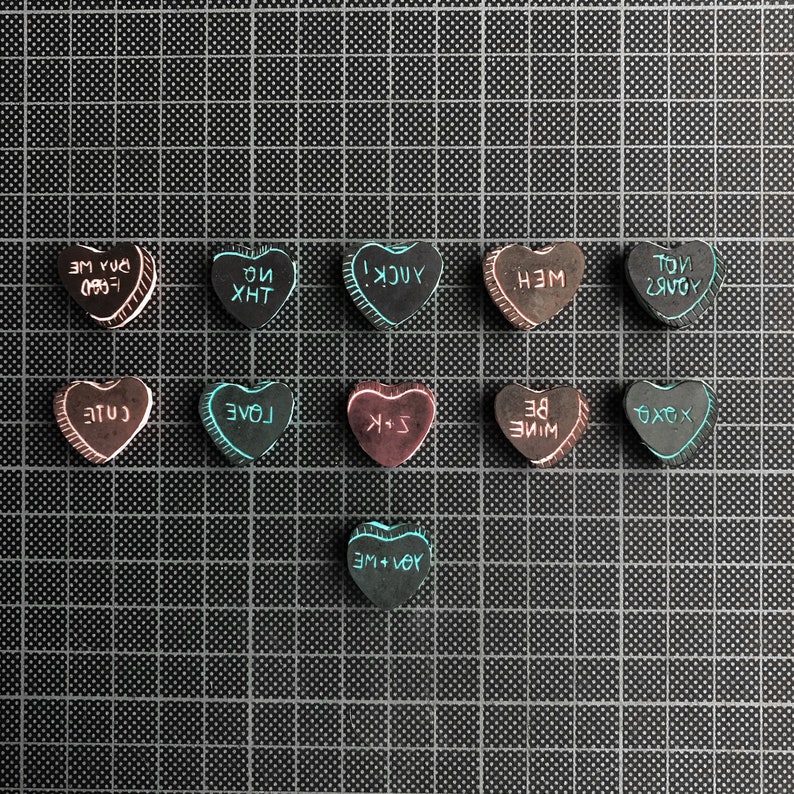 May include: A collection of 12 heart-shaped candy conversation hearts with various messages written on them, including "YOU + ME", "LOVE", "BE MINE", "XOXO", "YUCK", "THX", "CUTE", "TOU + ME", "YOU'RE", and "TOU + ME". The hearts are arranged in three rows of four on a black grid background.