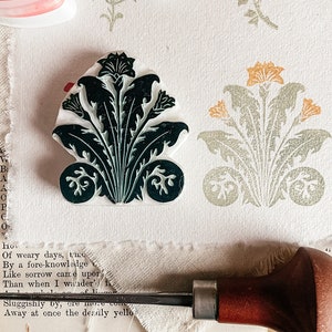May include: A close-up of a black and green floral stamp with a wooden handle and ink pads in the background. The ink pads are labeled "Drop Magic" and are orange, pink, and green.