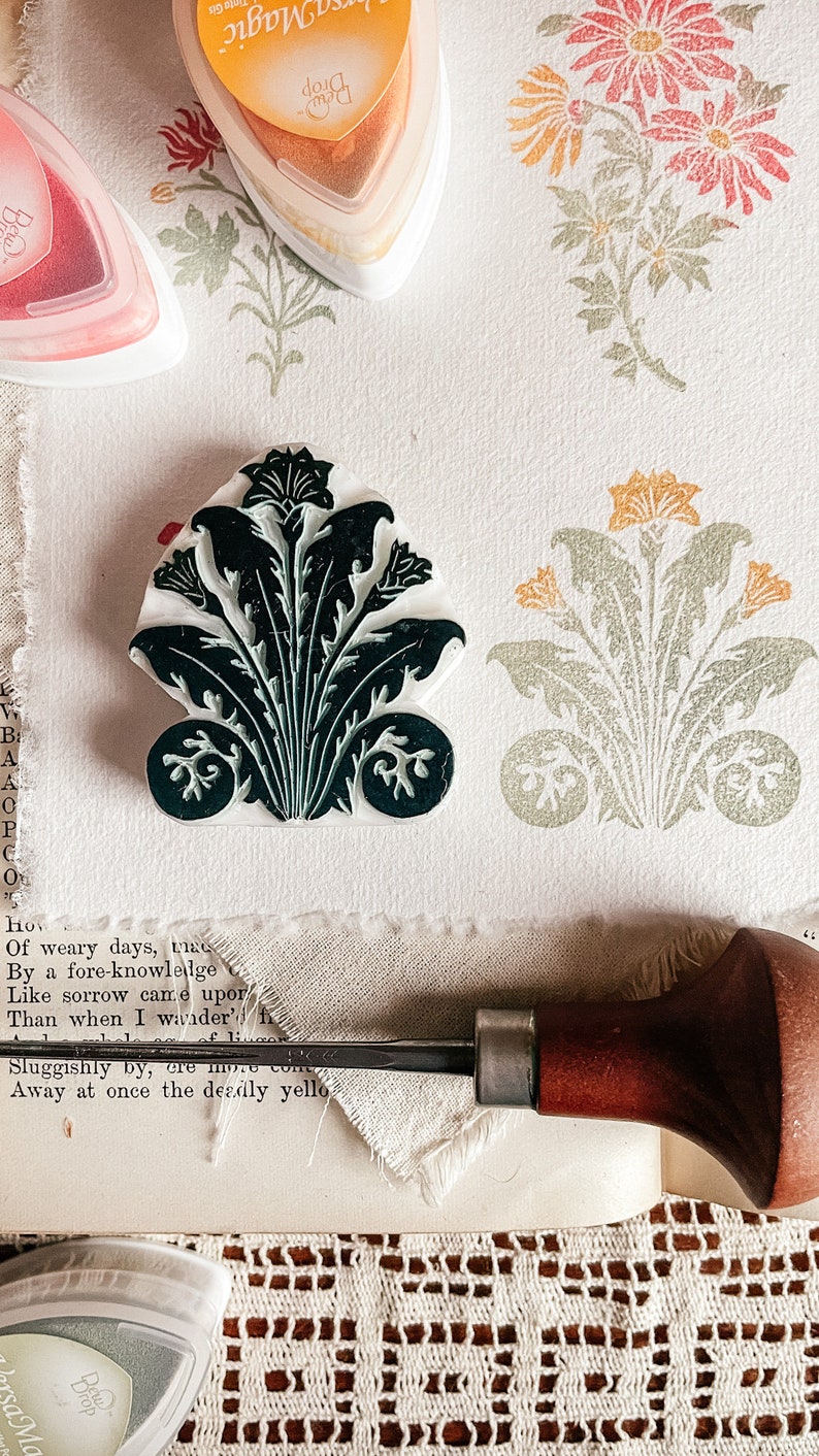 May include: A close-up of a black and green floral stamp with a wooden handle and ink pads in the background. The ink pads are labeled "Drop Magic" and are orange, pink, and green.