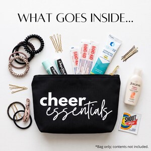 May include: A black zippered pouch with the white text "cheer essentials" printed on the front. Surrounding the pouch are various items including hair ties, bobby pins, lip balm, adhesive bandages, hydration powder, lotion, and a stain wipe.