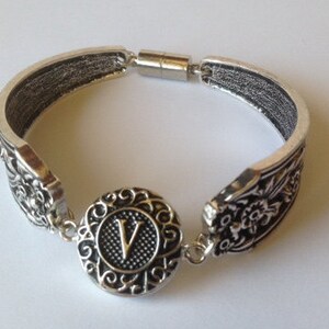 May include: Silver bracelet with a floral design and a magnetic clasp. The bracelet features a round charm with the letter "V" in the center.