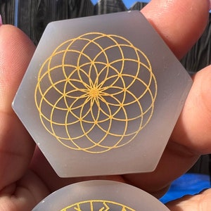 May include: Two white agate coasters with gold geometric designs. One coaster features a flower of life pattern, the other has a compass with runes around the edge.