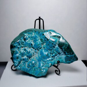 May include: A large, irregularly shaped piece of blue and green chrysocolla with a bubbly texture. The stone is displayed on a black metal stand.