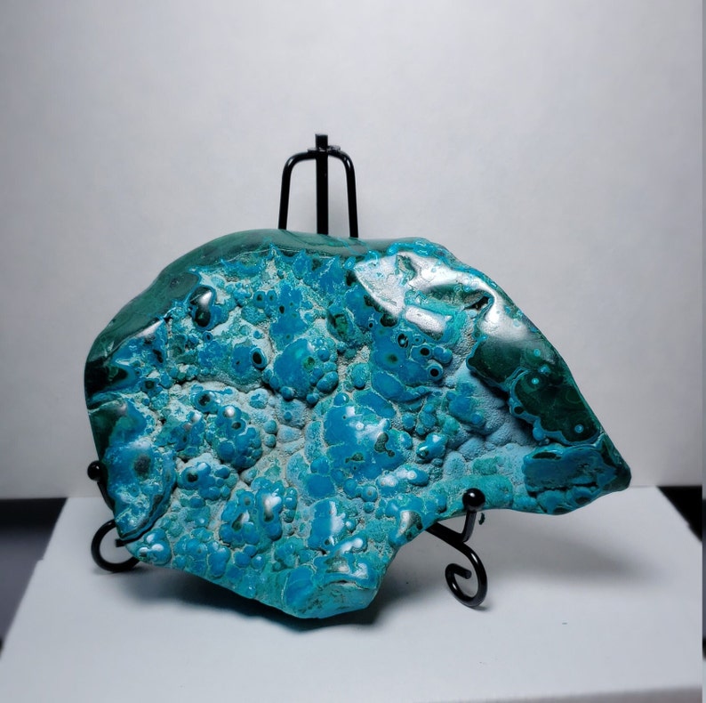 May include: A large, irregularly shaped piece of blue and green chrysocolla with a bubbly texture. The stone is displayed on a black metal stand.