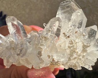 May include: A cluster of clear quartz crystals with a rough, natural texture. The crystals are clustered together in a random formation, creating a unique and interesting display.