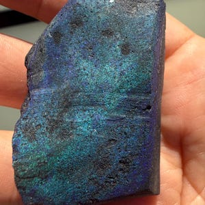 May include: A raw, irregularly shaped mineral specimen featuring a deep blue and iridescent teal surface. The texture is rough and porous with dark, matte black inclusions scattered across the face, creating a high-contrast, multi-tonal appearance.