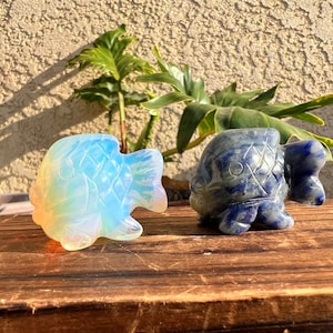 May include: Two carved fish figurines, one made of iridescent opalite and the other of blue lapis lazuli. The opalite fish is facing left and the lapis lazuli fish is facing right.