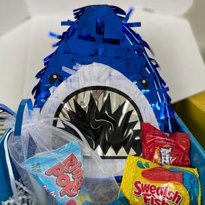 May include: A blue shark-shaped piñata with a white mouth and black teeth, accompanied by a white organza bag containing a blue Ring Pop, a blue Airheads Blue Raspberry candy bar, a yellow Swedish Fish Mini bag, and a red Twizzlers candy package.