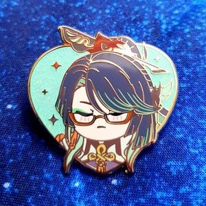 May include: A heart-shaped enamel pin featuring a cartoon character with green hair, wearing glasses and a gold and purple outfit. The character is surrounded by a light blue background with gold stars.