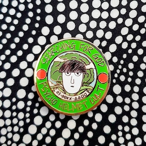May include: A green and gold enamel pin featuring a cartoon character wearing a helmet. The pin reads "Searching for God Psycho Helmet Cult 03-00XX-4000".