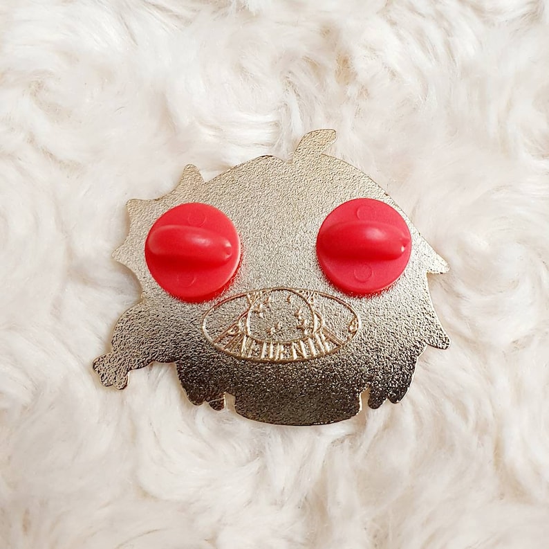 May include: A gold-coloured metal pin featuring a cartoon character design. The character has red eyes and a red tongue. The pin has a safety clasp on the back.