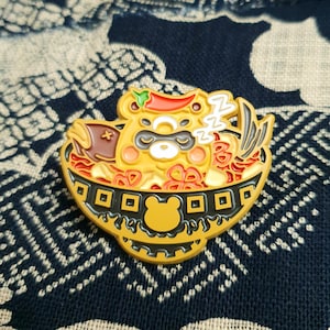 May include: A gold-coloured enamel pin depicting a cartoon animal with a red chilli pepper on its head, sleeping in a bowl of ramen noodles. The animal has black eyes and a white nose. The bowl is decorated with a black rim and a pattern of squares.