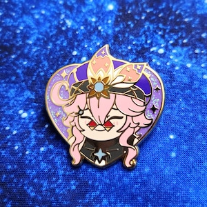 Può includere: Spilla smaltata a forma di cuore con un personaggio dei cartoni animati con capelli rosa, occhi rossi e un abito nero. La spilla ha un bordo dorato ed è decorata con accenti viola, rosa e blu. Il personaggio sorride.